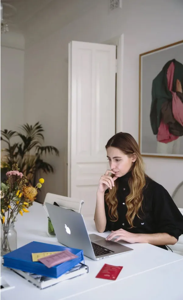 Young woman with long hair in a black shirt working on a laptop at a white desk with folders and flowers nearby.