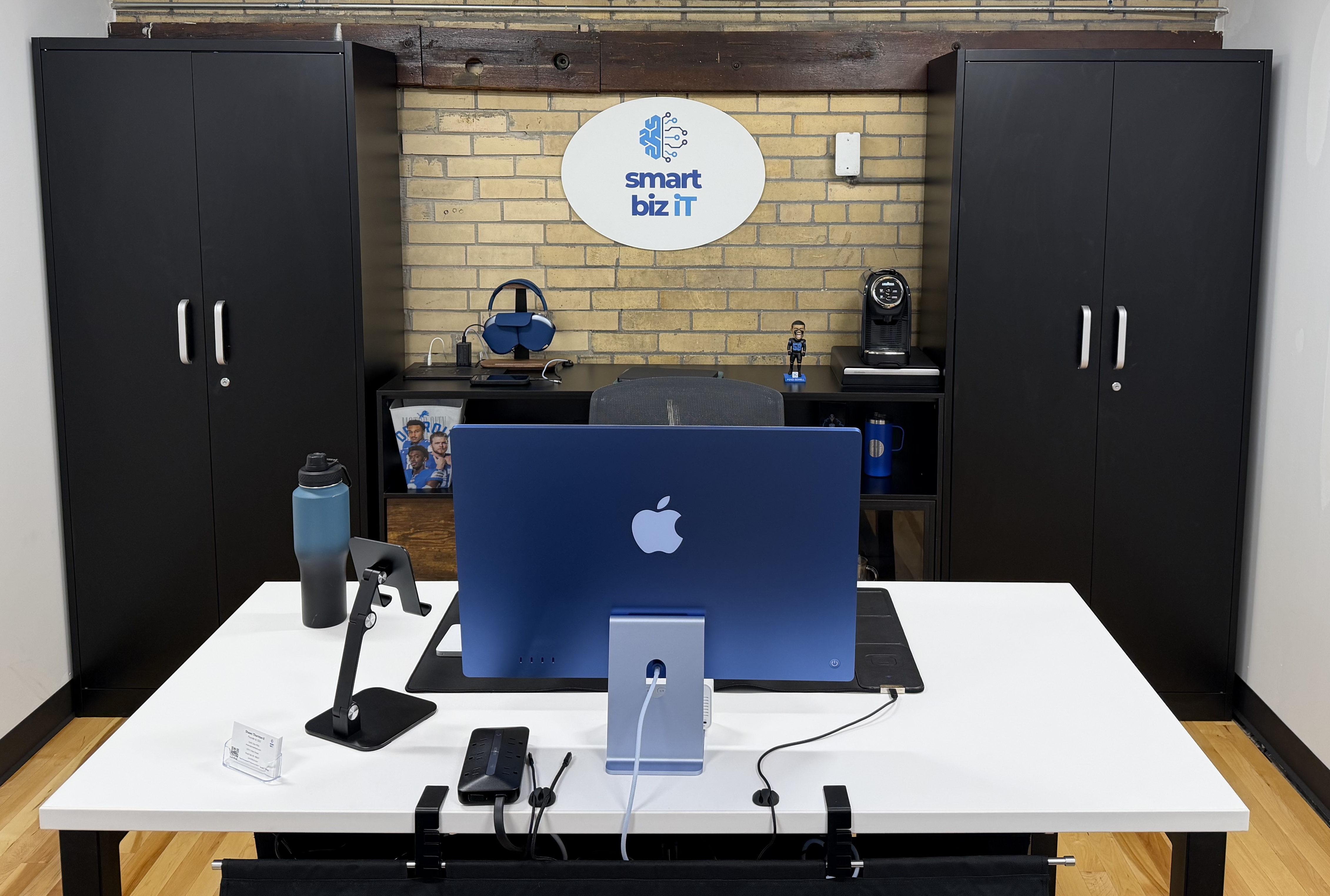 Modern office desk with a blue Apple iMac, water bottle, phone stand, and black cabinets against a brick wall with a Smart Biz IT sign.