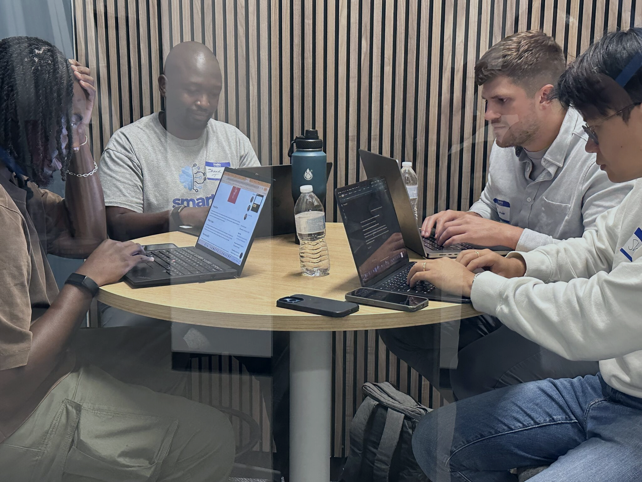 Four people sitting around a round wooden table working on laptops, with water bottles and smartphones on the table, in a room with vertical wooden paneling.