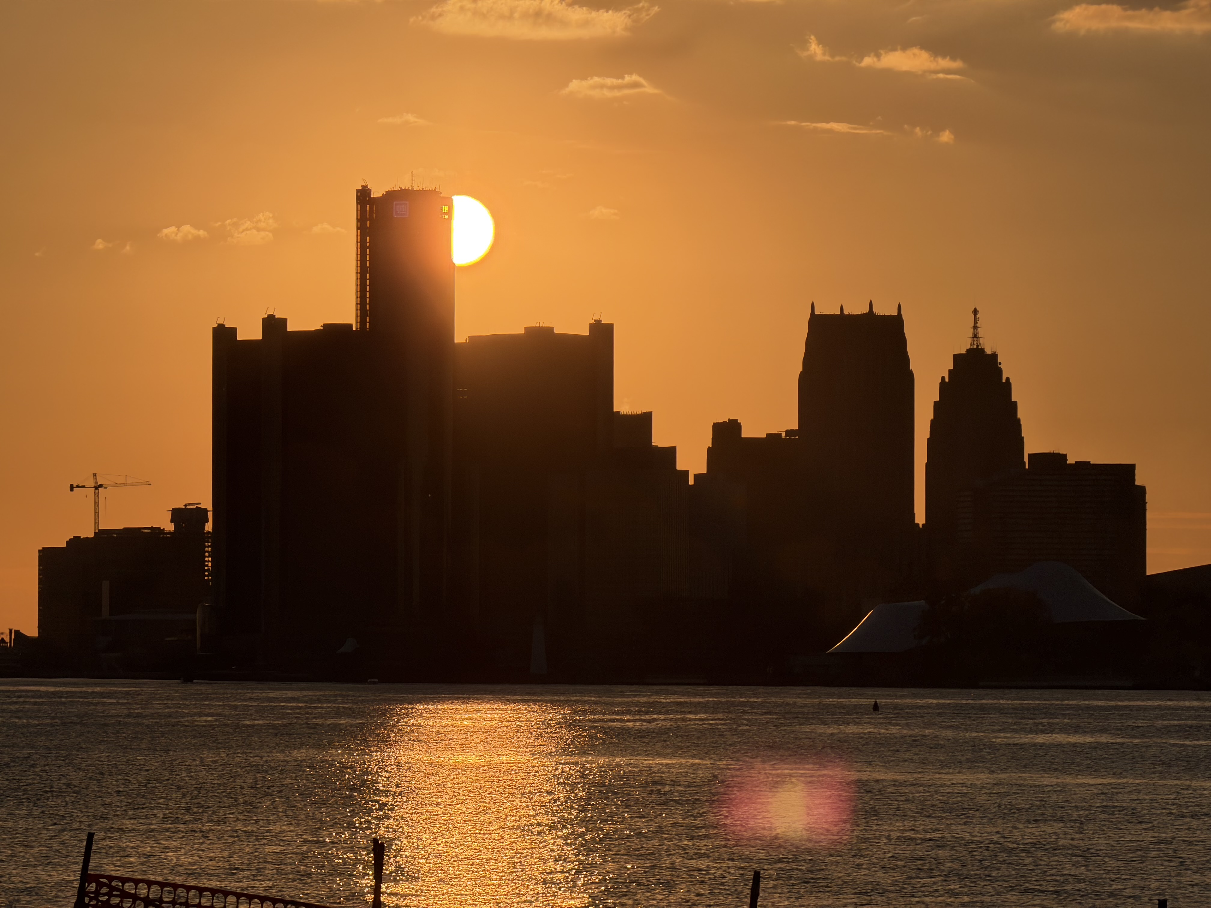 Silhouette of Detroit skyline at sunset with the sun partially visible behind a tall building and its reflection on the water.