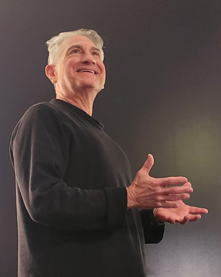 Smiling older man with gray hair wearing a black long-sleeve shirt gesturing with his hands against a dark background.