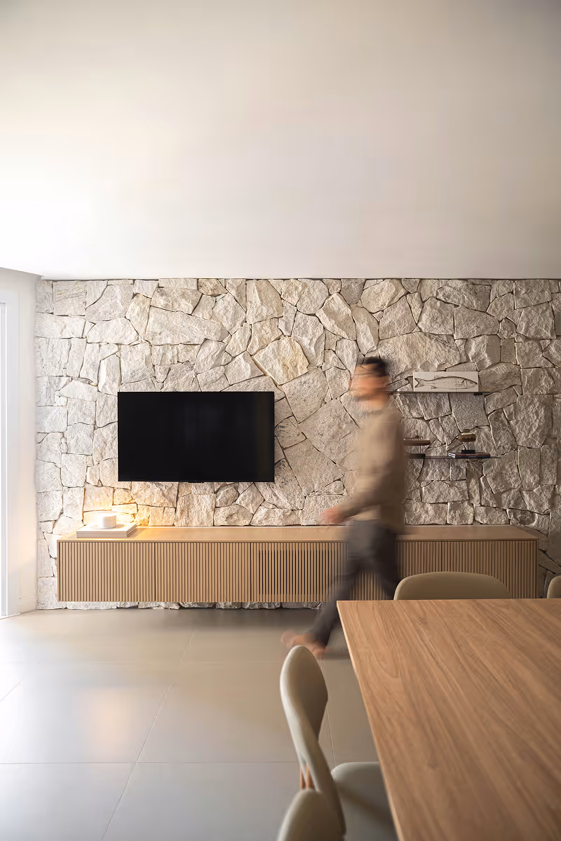 Modern living room with stone wall, wall-mounted TV, wooden cabinet, and blurred person walking.