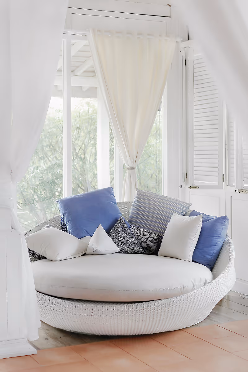 Round white wicker chair with white and blue decorative pillows near large window with white curtains.
