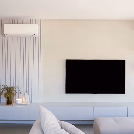 Modern living room with a wall-mounted flat-screen TV, white air conditioner, light-colored sofa, and minimalist decor including a potted plant and candles.
