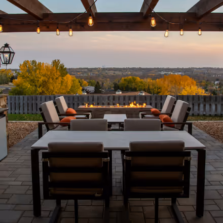 Outdoor patio with dining tables and cushioned chairs under string lights, overlooking a fire pit and scenic autumn trees.