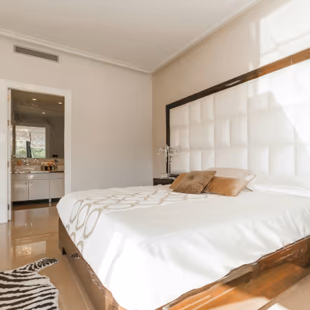 Bright bedroom with a large bed featuring a white tufted headboard, beige walls, and an open doorway leading to a bathroom.