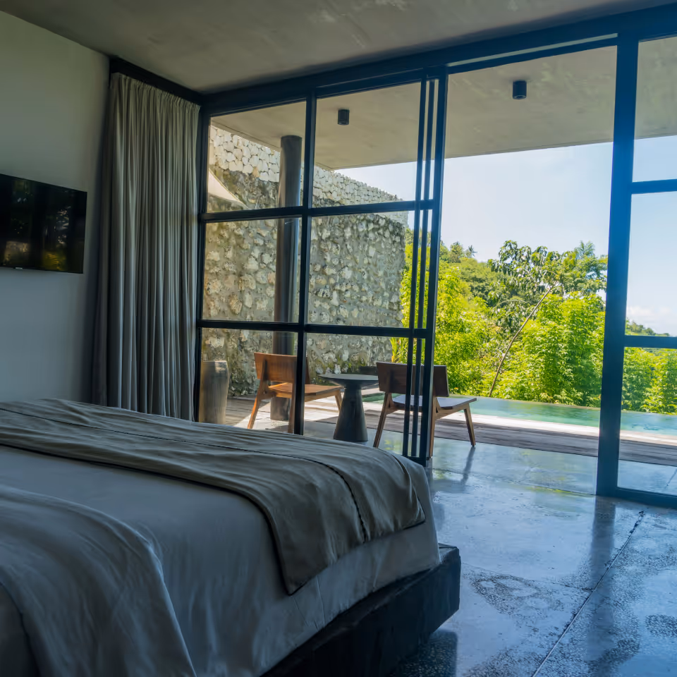 Modern bedroom with a large bed facing floor-to-ceiling windows that open to a patio with wooden chairs and a small table overlooking lush greenery.