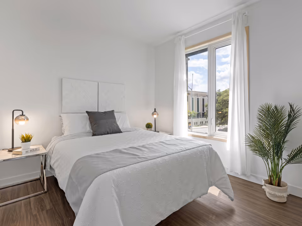 Bright bedroom with a neatly made bed with white and gray bedding, a side table with a lamp and plant, a large window with white curtains, and a potted green plant on wooden floor.