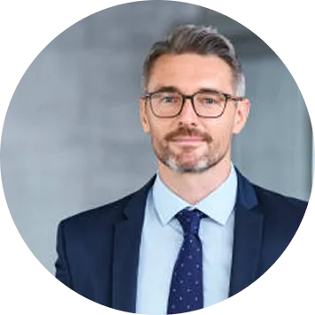 Man with glasses and a beard wearing a suit and tie in front of a blurred gray background.