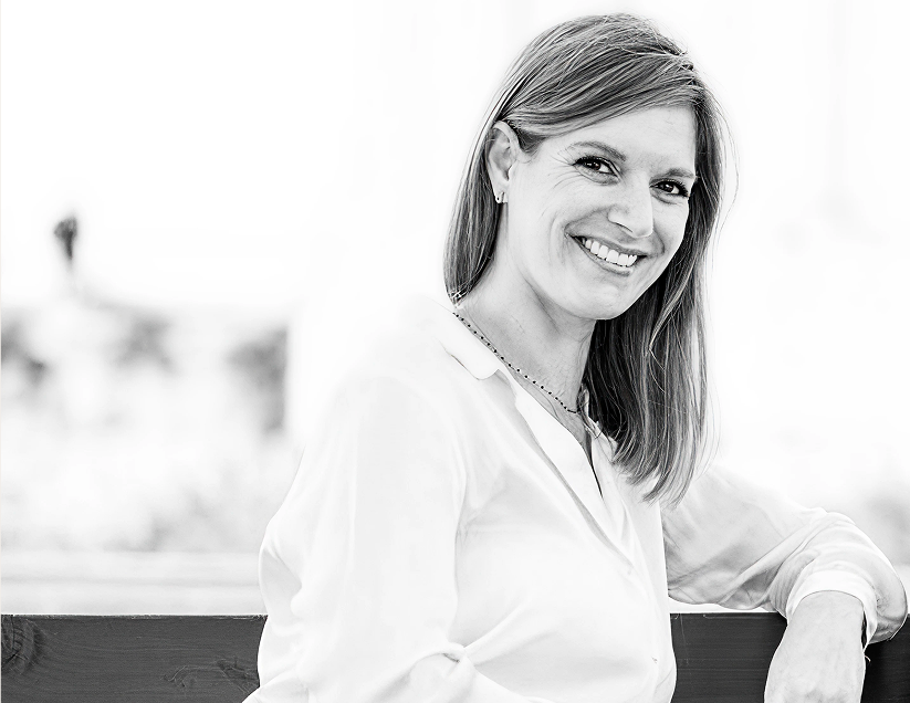 Smiling woman with straight hair wearing a white shirt, sitting and leaning on a wooden surface.