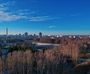 City skyline with mixed residential and industrial buildings under a clear blue sky, with leafless trees and a small body of water in the foreground.