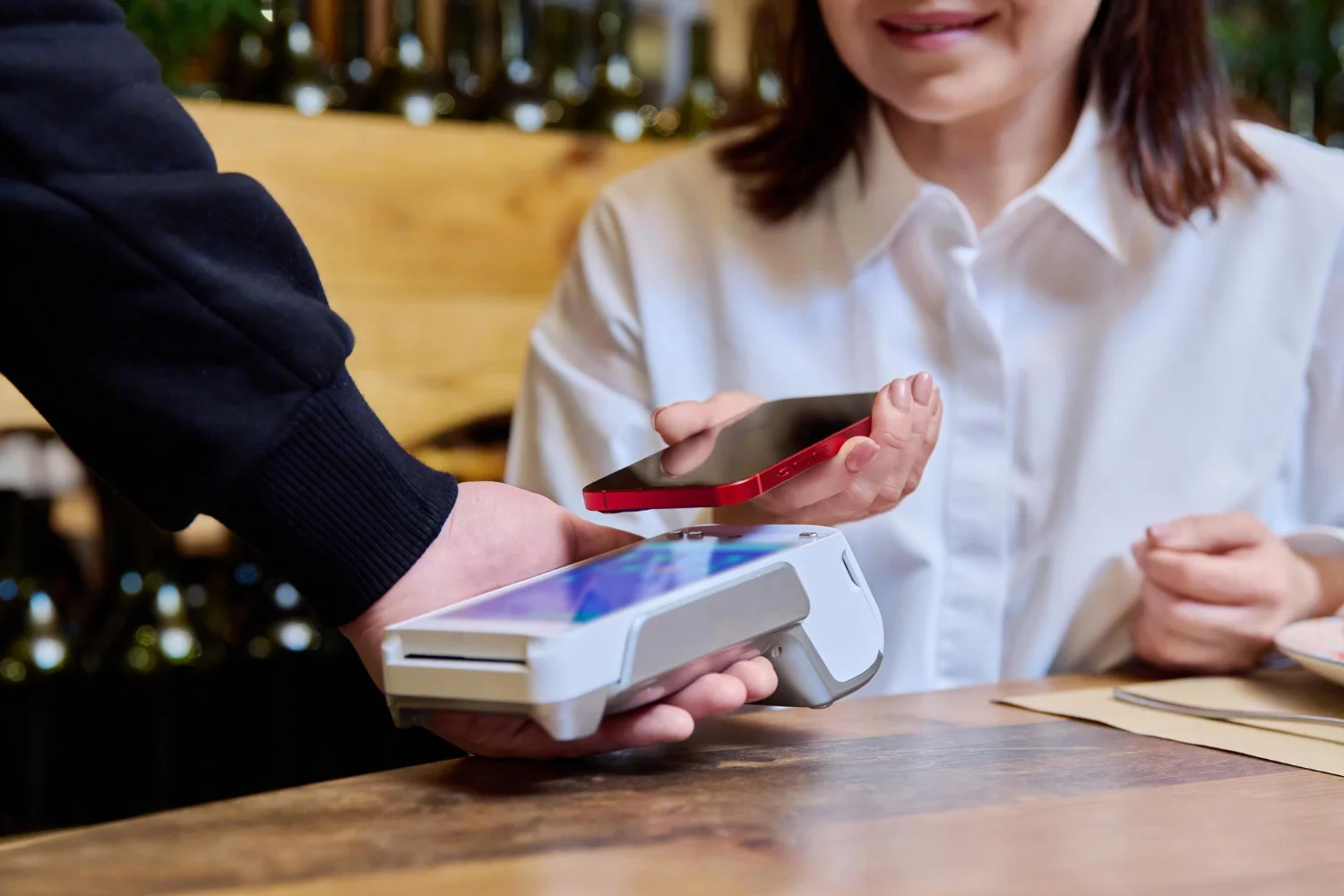 Person in white shirt making a contactless payment with a red smartphone on a white card reader at a wooden table.