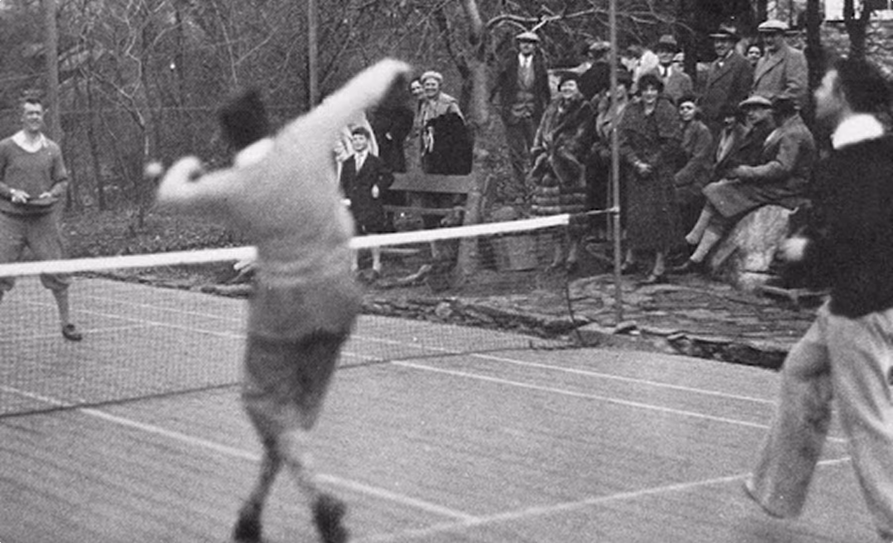 Black and white photo of people playing tennis outdoors with spectators in early 20th-century clothing watching behind the net.