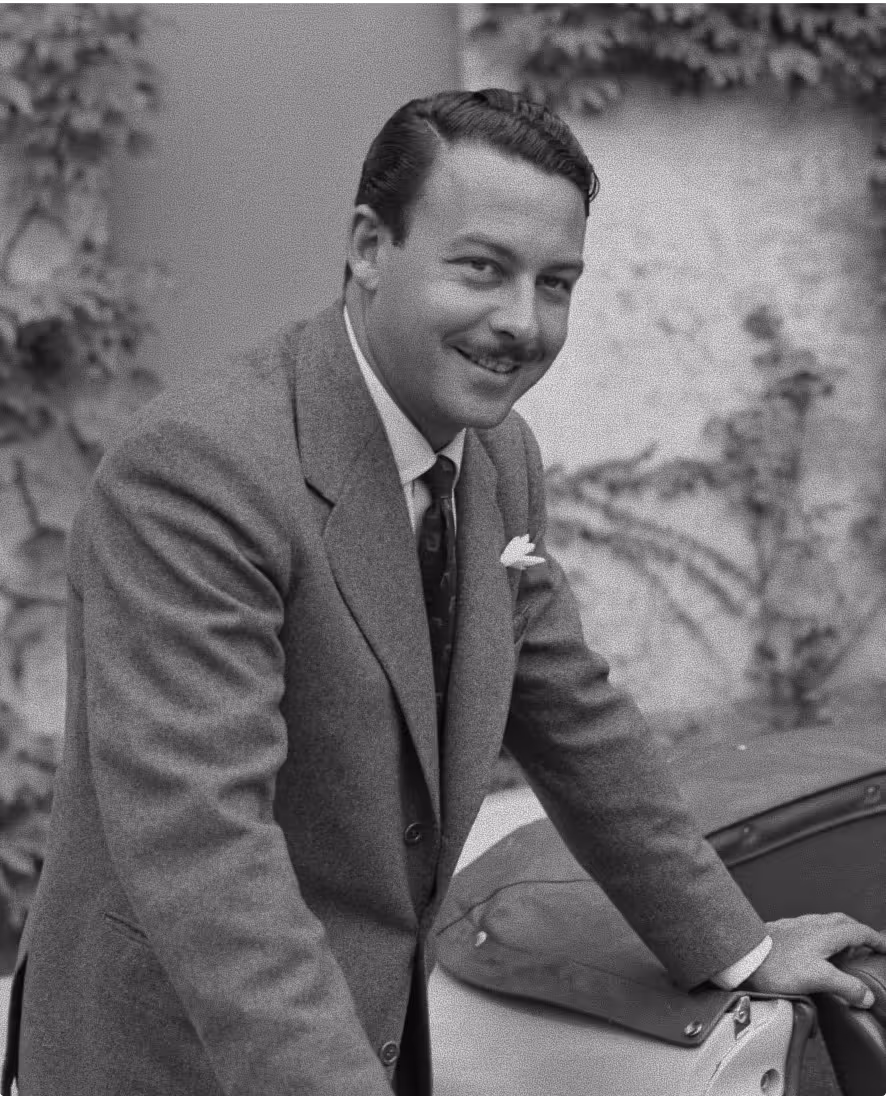 Black and white portrait of a smiling man in a suit leaning on a car door outdoors.