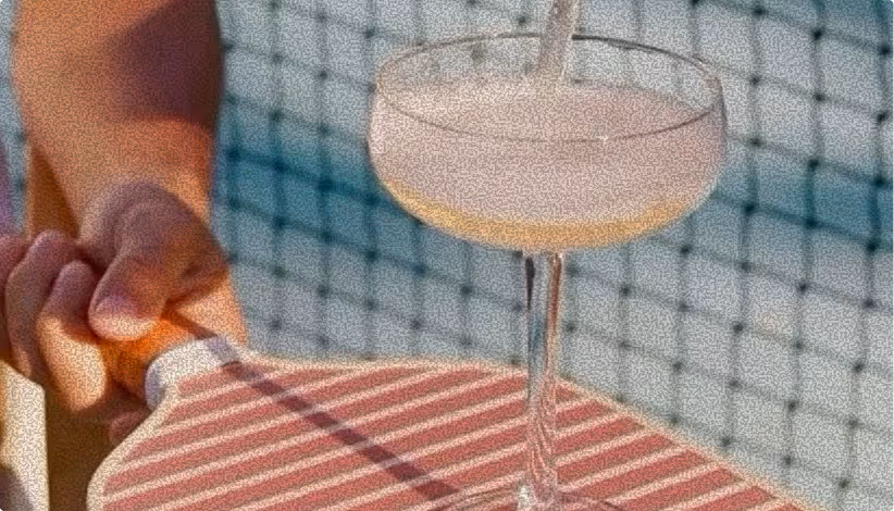 Close-up of a hand holding a wooden tennis racket with a cocktail glass placed on the racket's strings in front of a tennis net.