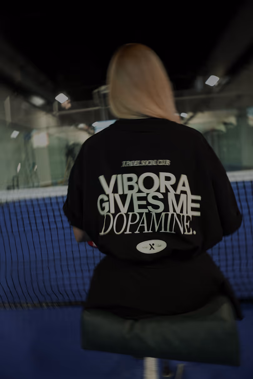 Person with light-colored hair sitting on a bench facing away, wearing a black shirt that reads 'VIBORA GIVES ME DOPAMINE' on the back in an indoor tennis court.