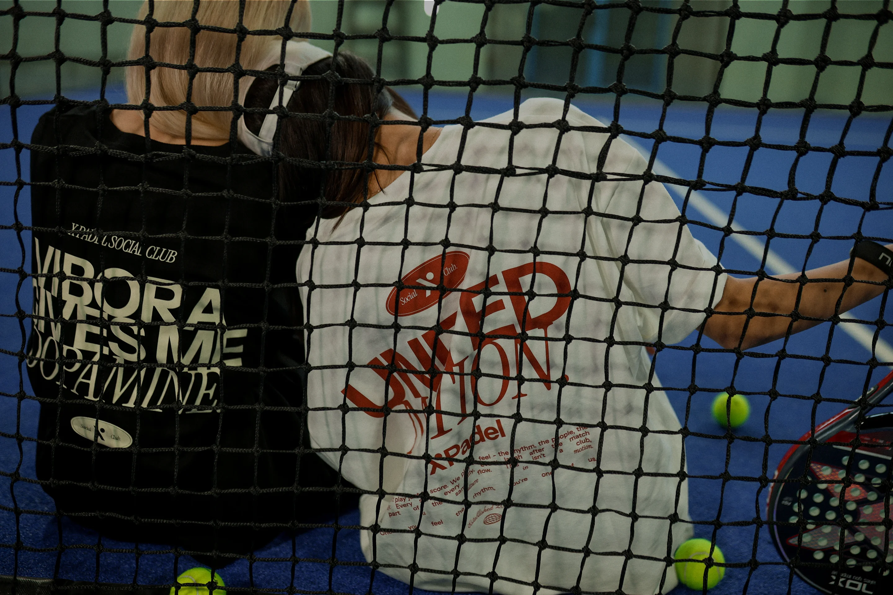 Two people sitting on a blue tennis court behind a black net, wearing black and white shirts with large printed text, surrounded by tennis balls and rackets.