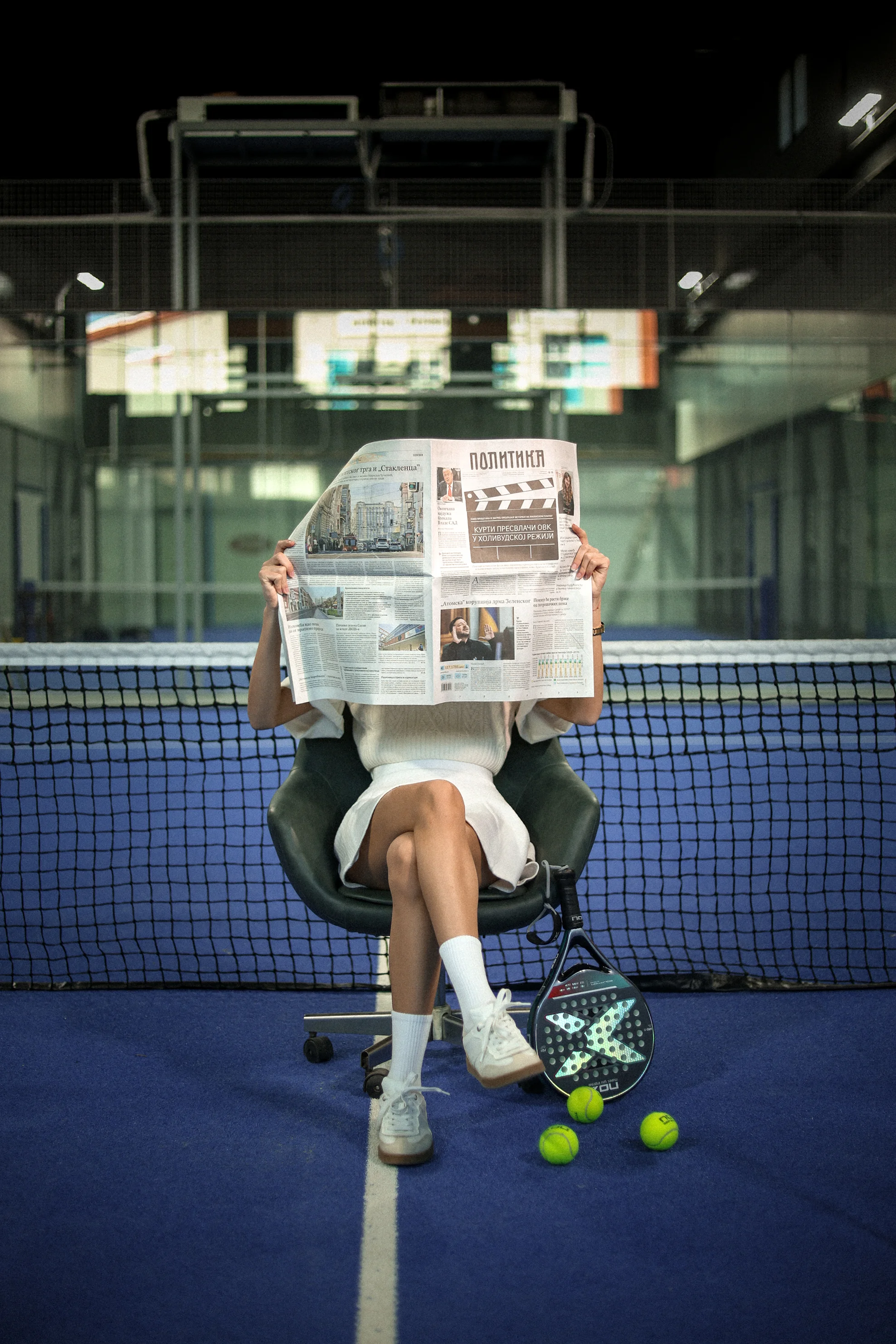 Person sitting on a chair on a blue paddle tennis court, reading a newspaper, with a paddle racket and tennis balls on the ground.