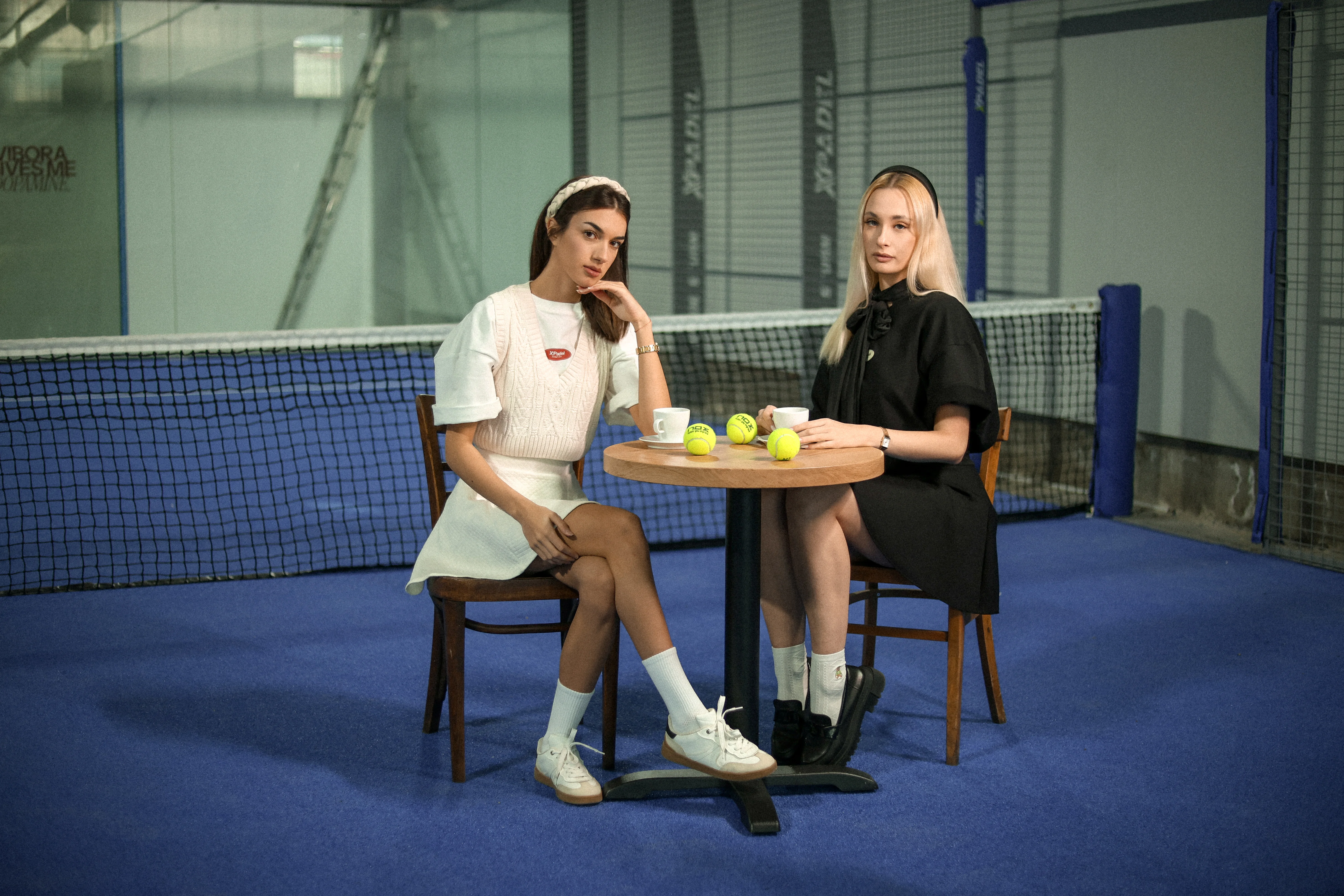 Two women sitting at a round table inside a tennis court with tennis balls and coffee cups on the table.
