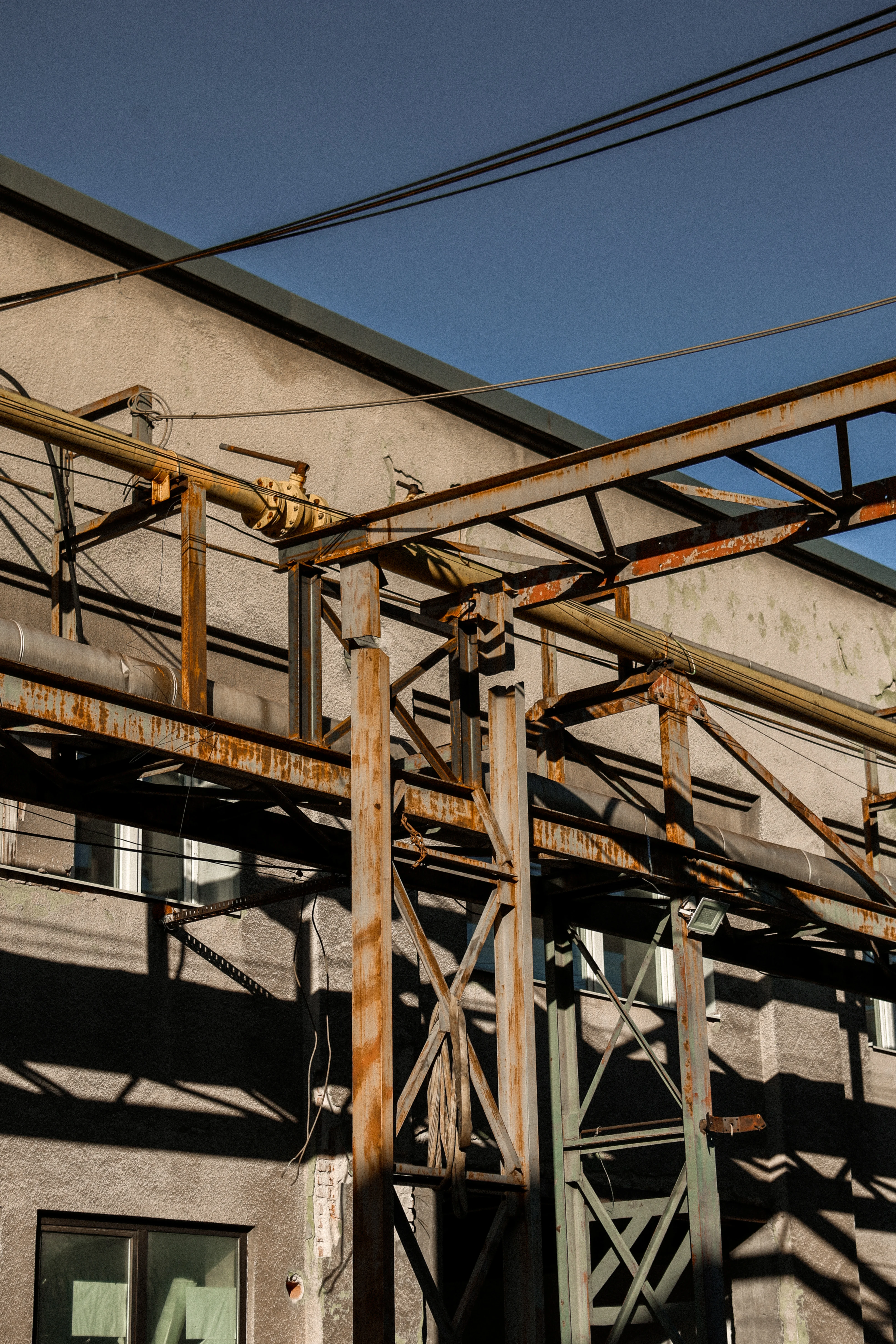 Rusty metal framework and pipes attached to an industrial building with windows under clear blue sky.