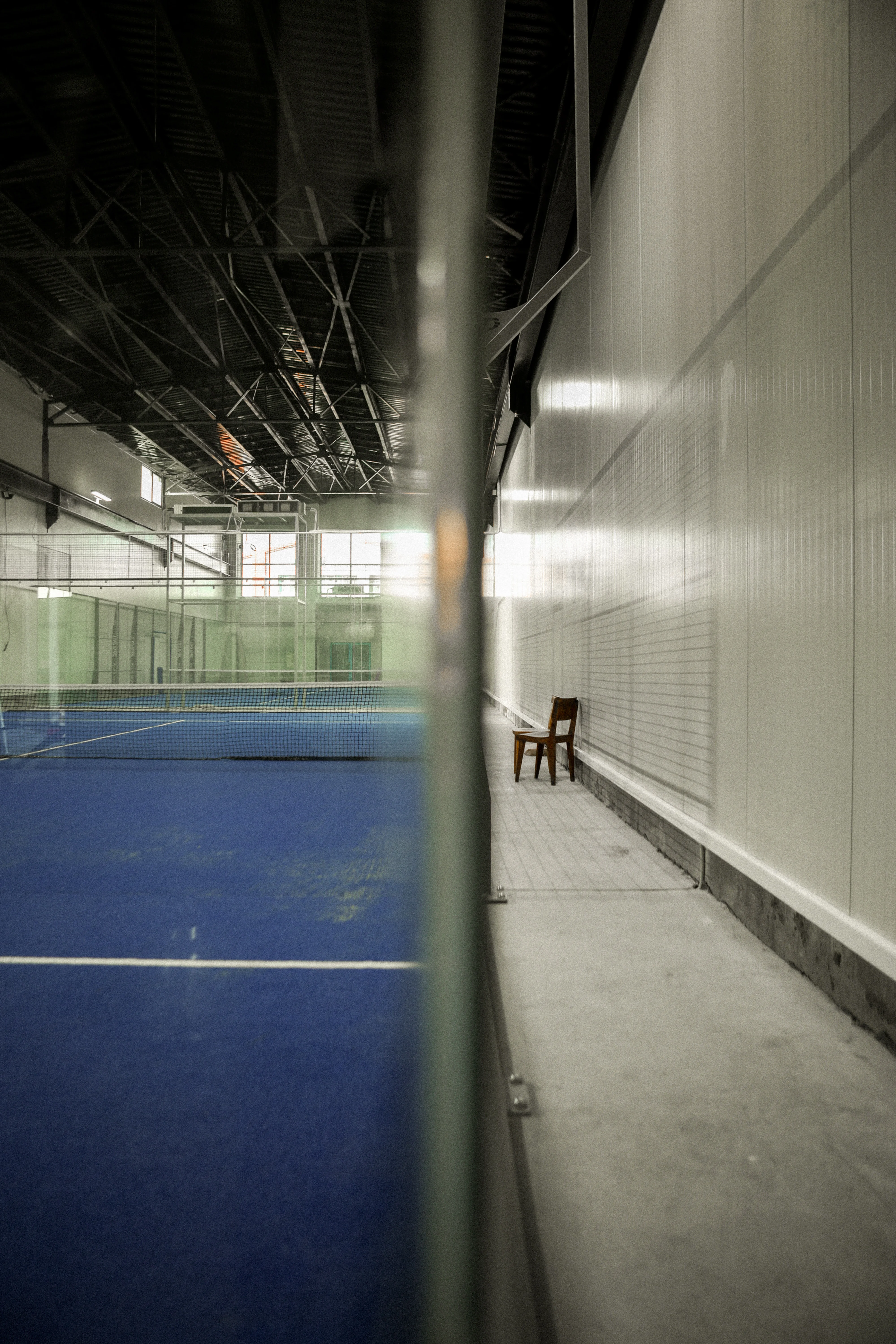Indoor padel court with a blue playing surface and a wooden chair placed against a white wall outside the court.