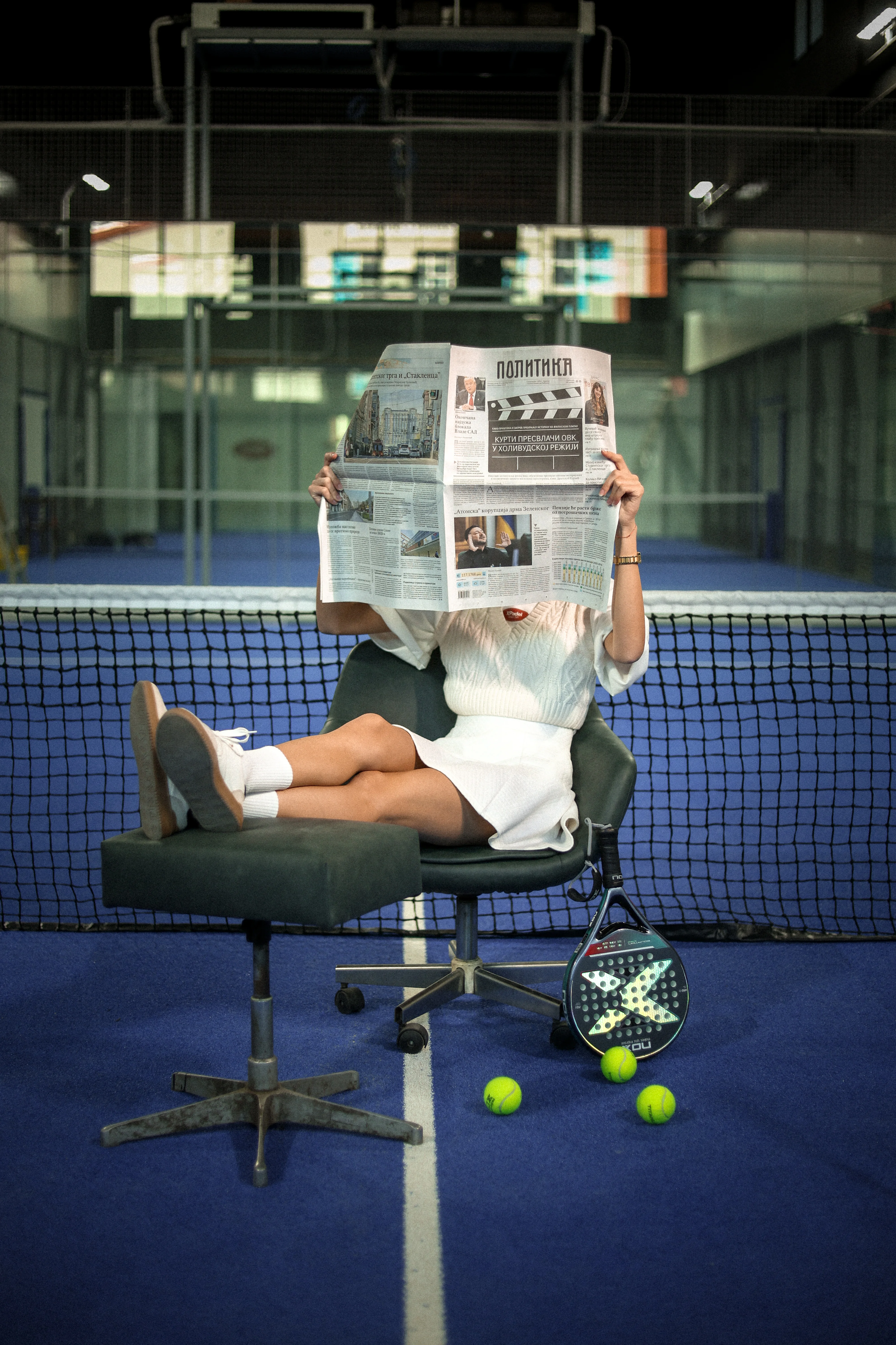 Person in white outfit sitting on office chair with feet on stool, reading a newspaper on an indoor tennis court with paddle and tennis balls nearby.