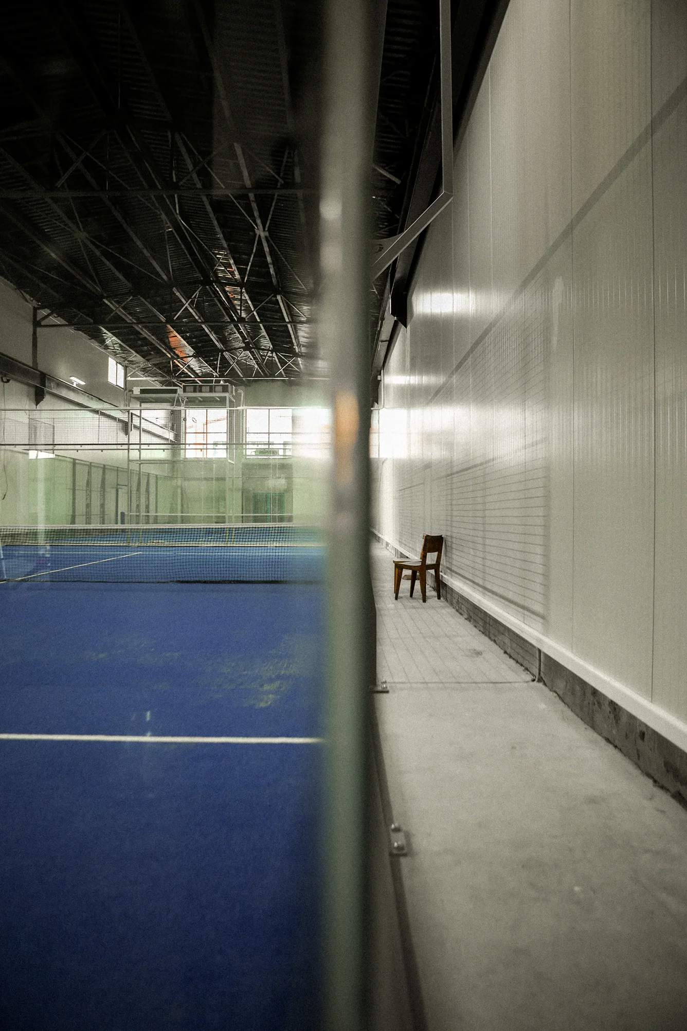 Indoor tennis court with blue surface and net, viewed through a glass barrier with a wooden chair against a white wall on the right.