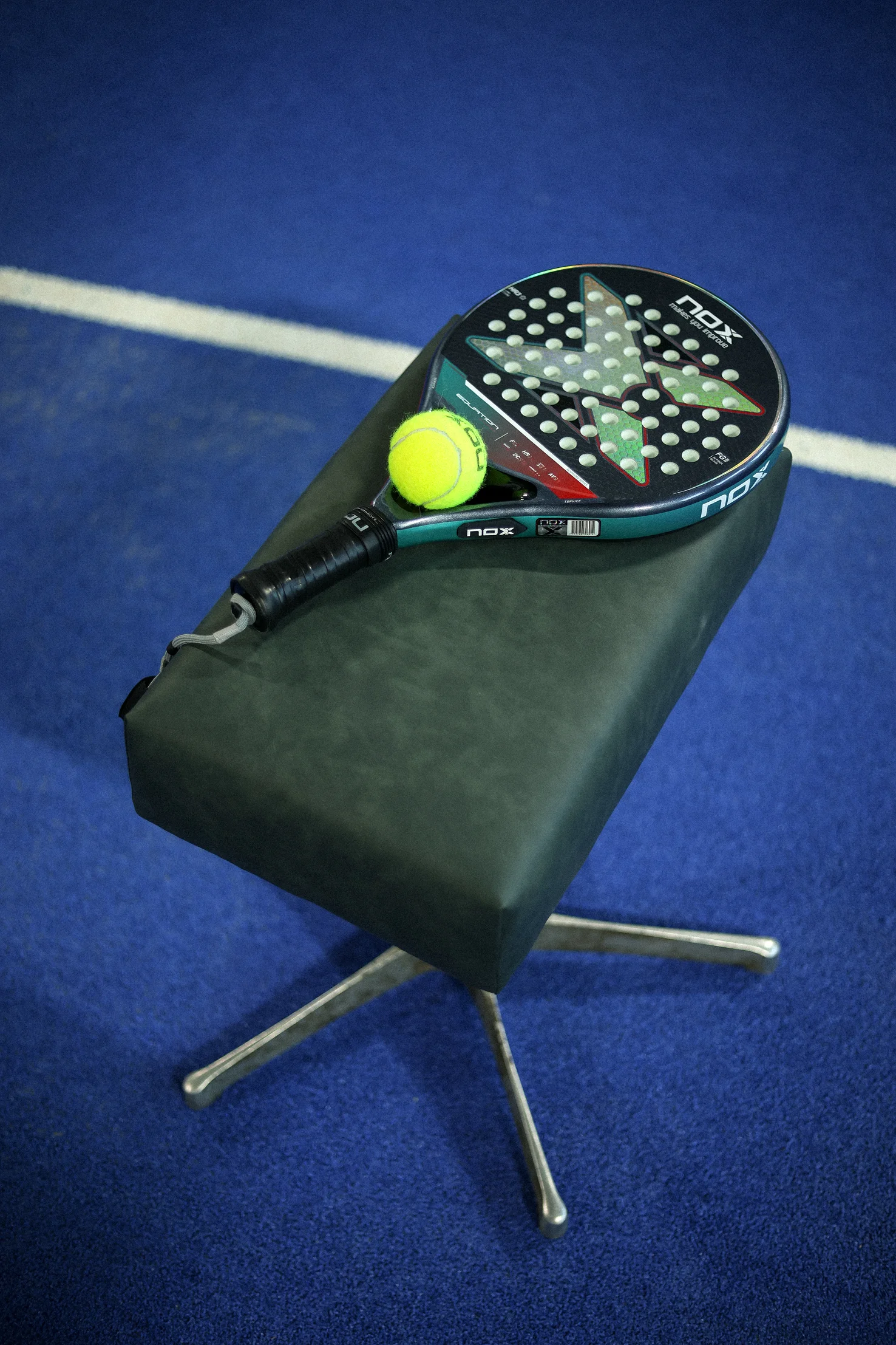 Black and turquoise NOX paddle racket with a bright yellow tennis ball resting on a green padded stool on a blue tennis court.