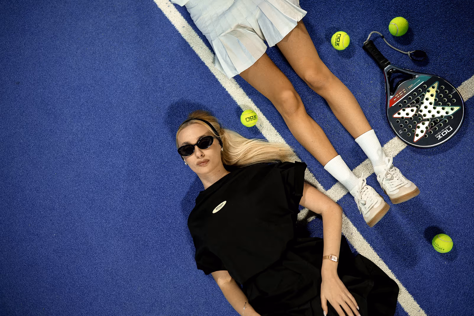 A woman in black sunglasses and dress lying on a blue sports court next to a person in white tennis skirt, white socks, white sneakers, tennis balls, and a paddle racket.