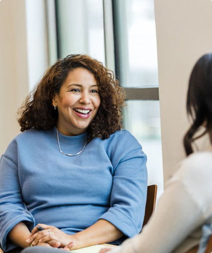 Smiling woman with curly hair in a blue sweater sitting and talking with another person by a window.