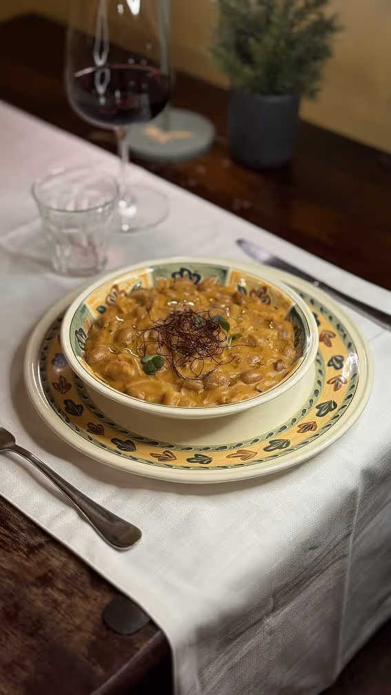 Piatto di pasta cremosa con fagioli, guarnita con erbe fresche e fili sottili rossi, servita su piatti decorati accanto a un bicchiere di vino rosso e un bicchiere d'acqua.