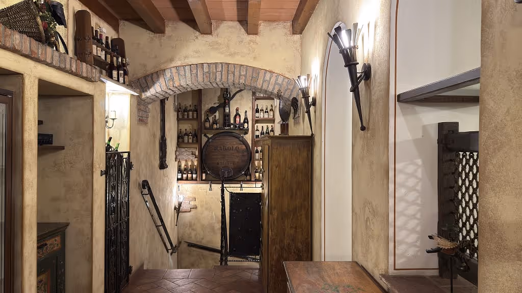 Interno rustico con pareti color crema, soffitto con travi in legno, lampade a muro e scaffali con bottiglie di vino, e botte di legno al centro.