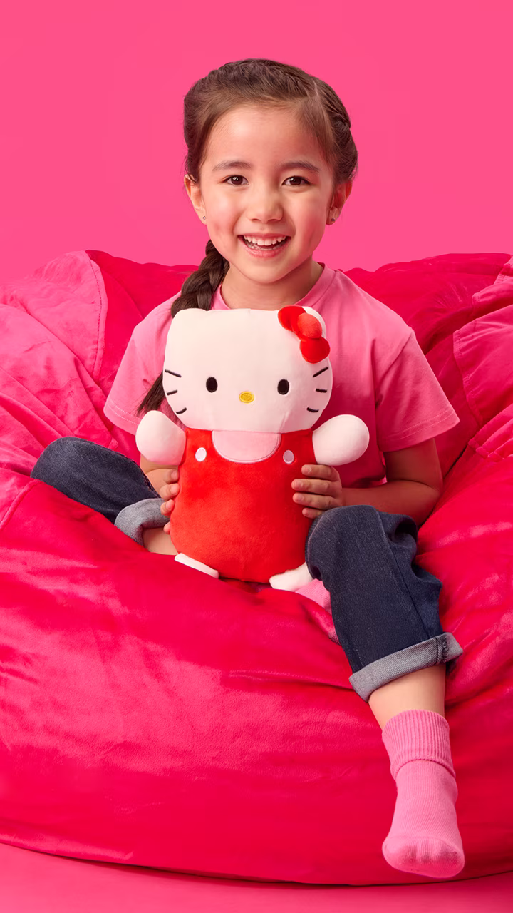 Child holding a HugMees Squishmallows plush character on a colorful studio set