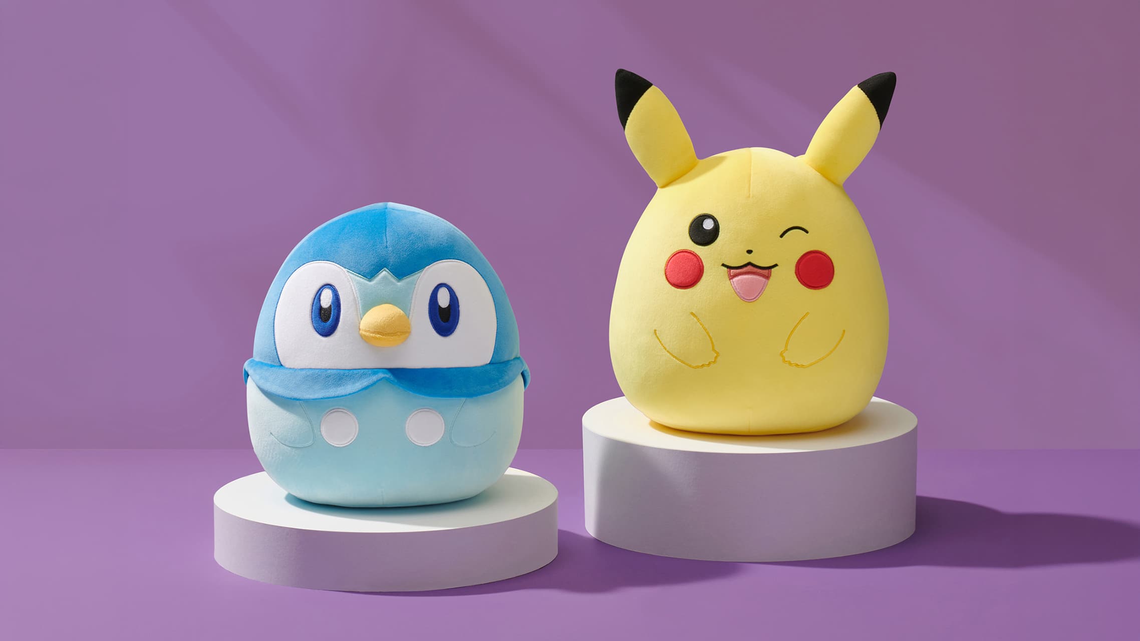 Piplup and Pikachu Pokémon plush toys displayed on pedestal stands against purple background