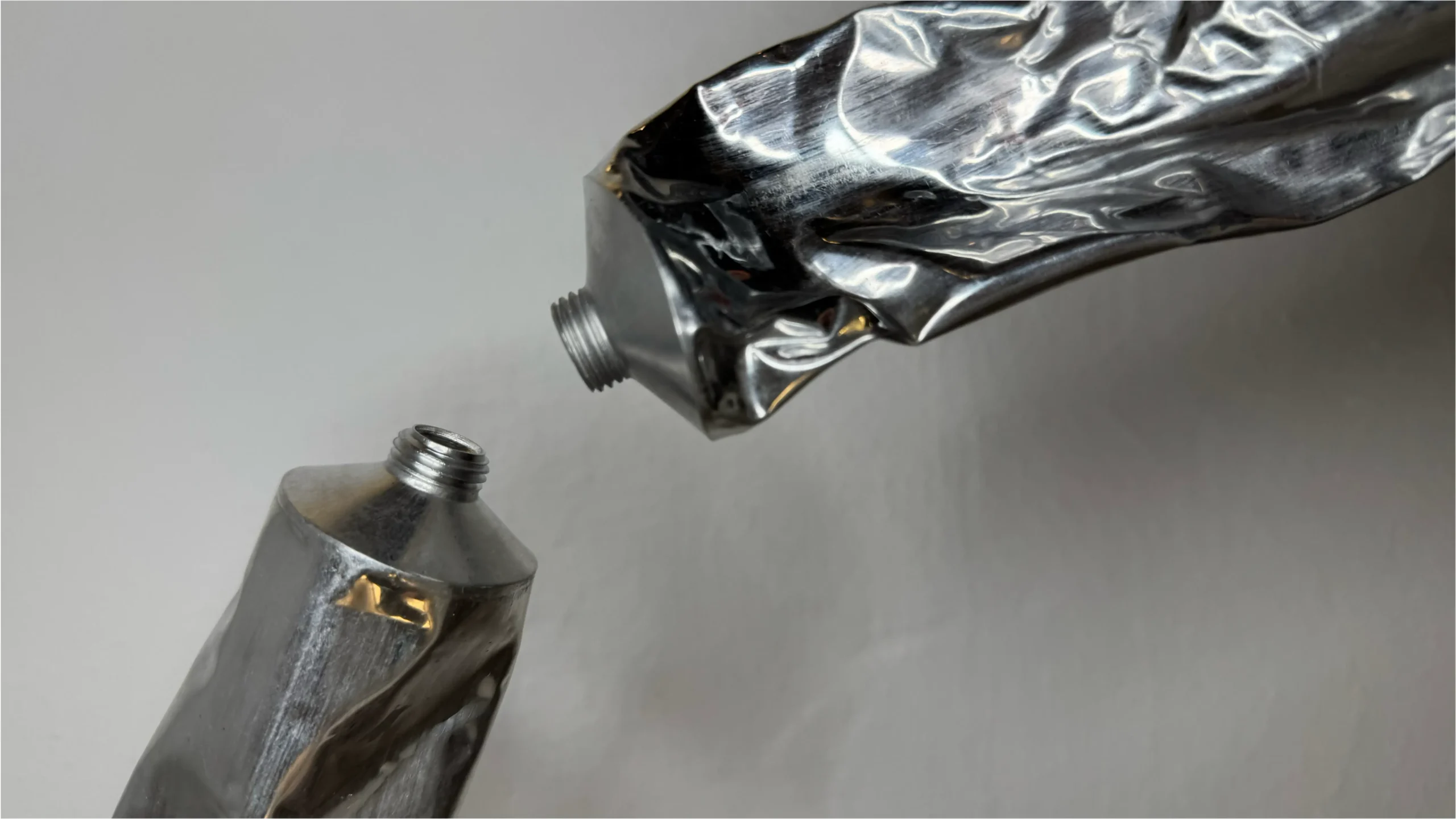 To sammenklemte metalltuber med åbne gevindede mundstykker mod en grå baggrund.