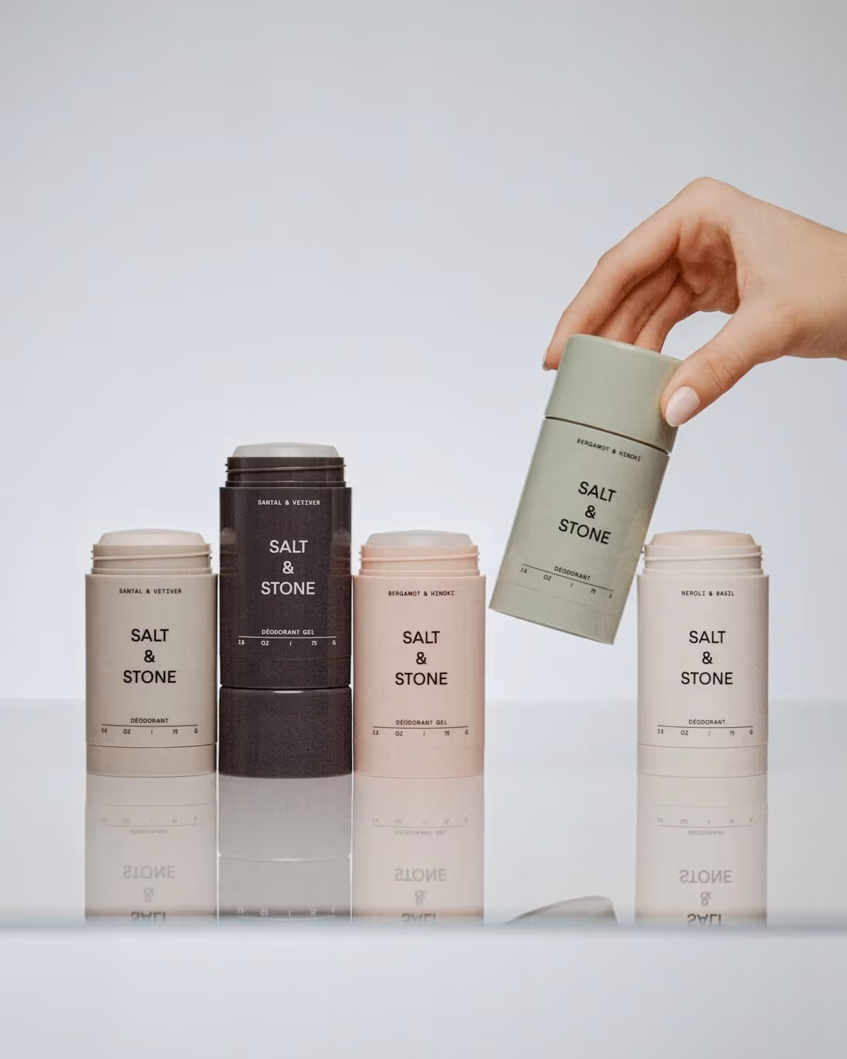 Fem forskellige Salt & Stone deodorantstick med labels i neutrale farver, hvor en hånd holder en grøn deodorant.