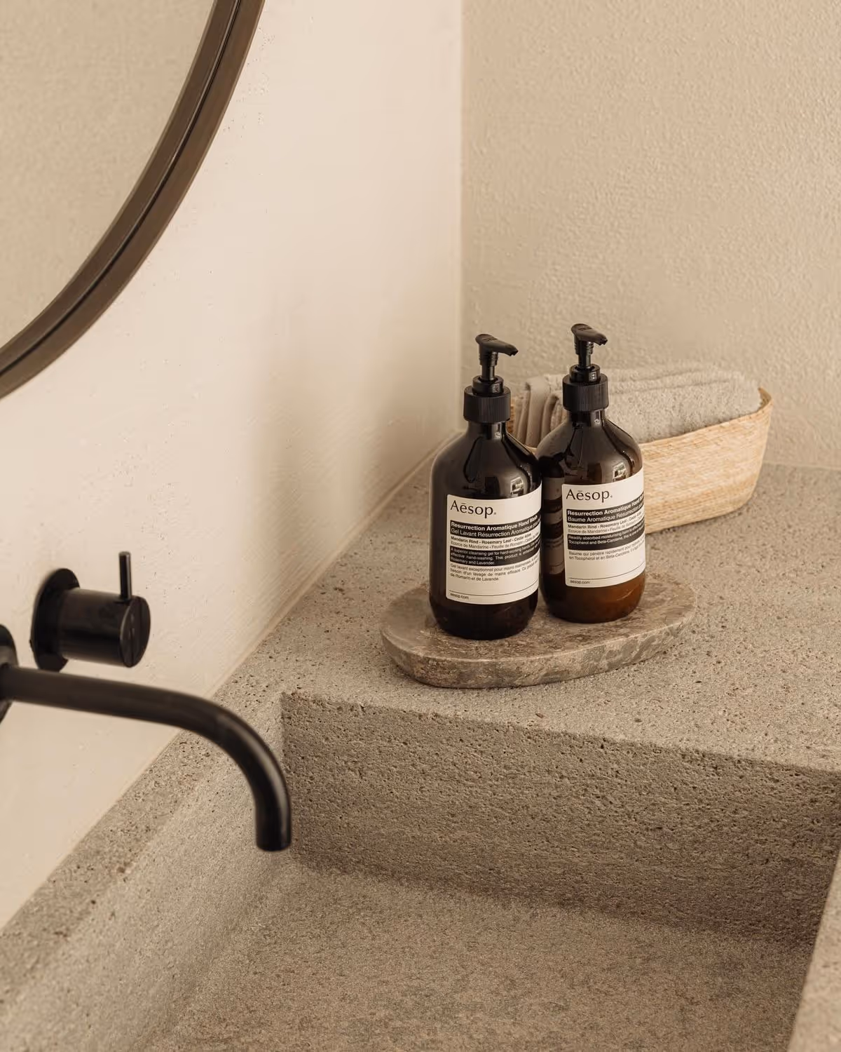 Toiletbord med to Aesop-flasker med pumpe ved siden af et fad og et sæt foldede håndklæder i en kurv.