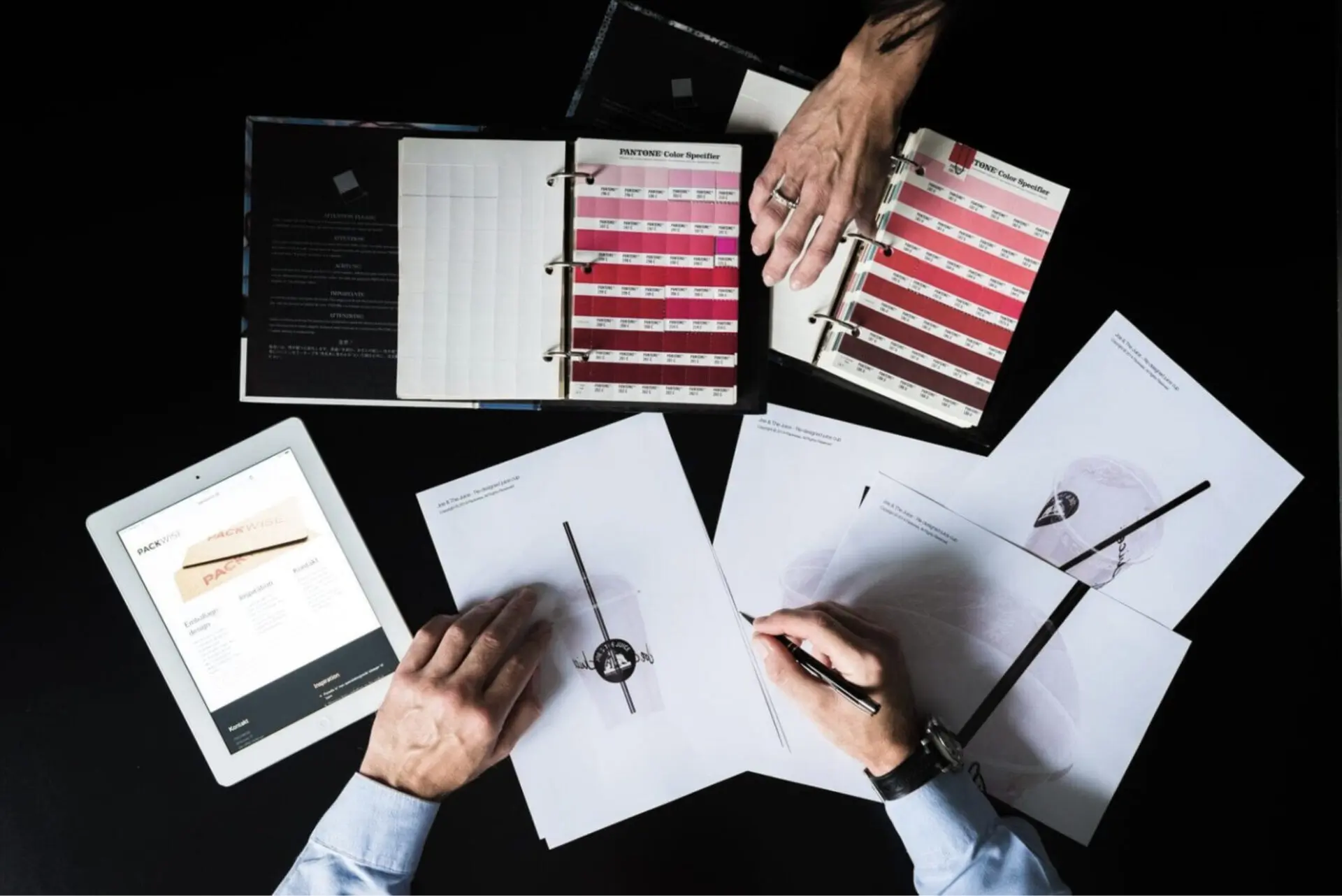 To hænder arbejder på emballagedesign med trykte dokumenter af plastikbægre, Pantone-farvekort og en tablet med emballagedesignside på en sort baggrund.