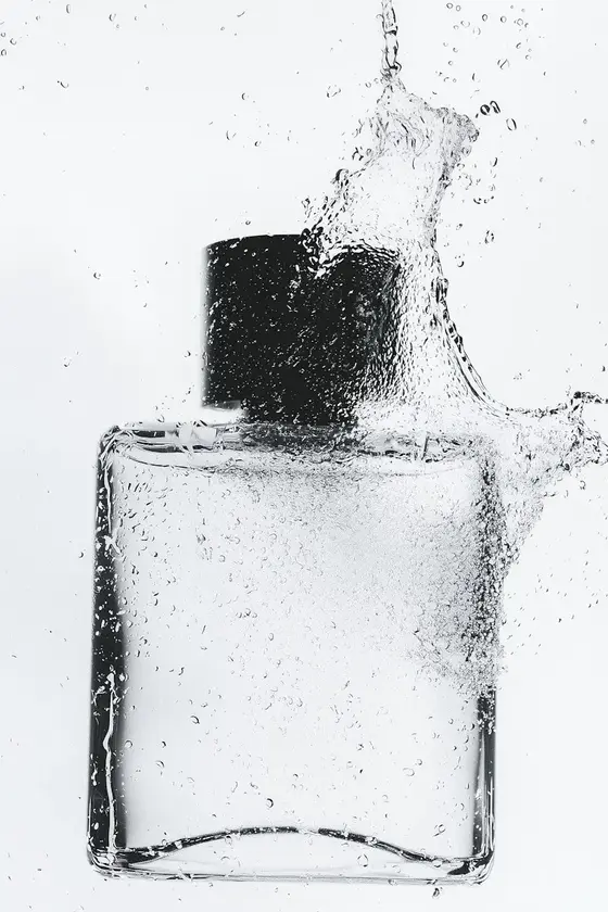 Glasflaske med sort hætte omfavnet af sprøjtende vanddråber på hvid baggrund.