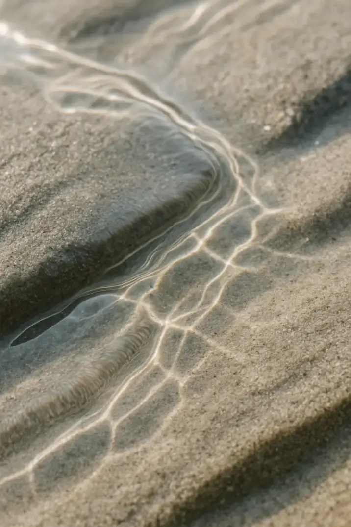 Detalje af sand med små bølger og lysrefleksioner fra vand.