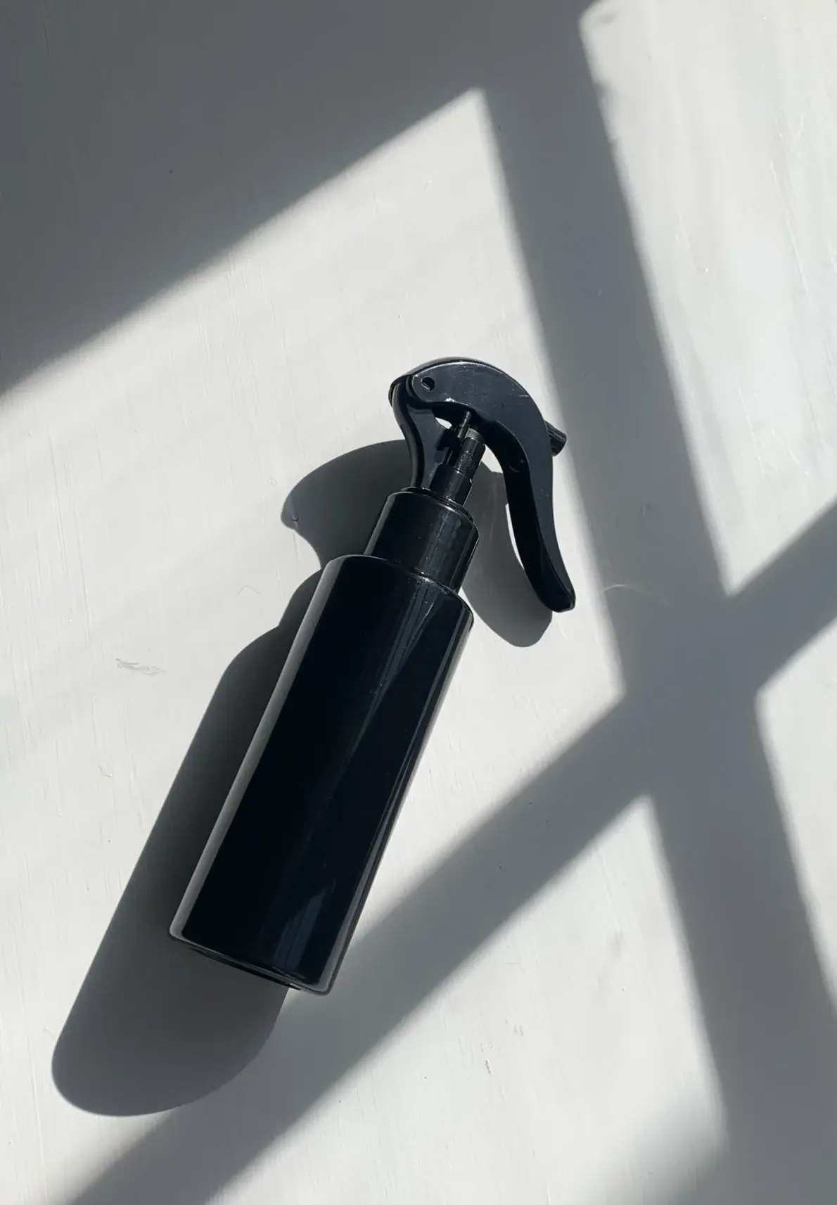 Sort sprayflaske på hvidt bord med skygger fra vindue.