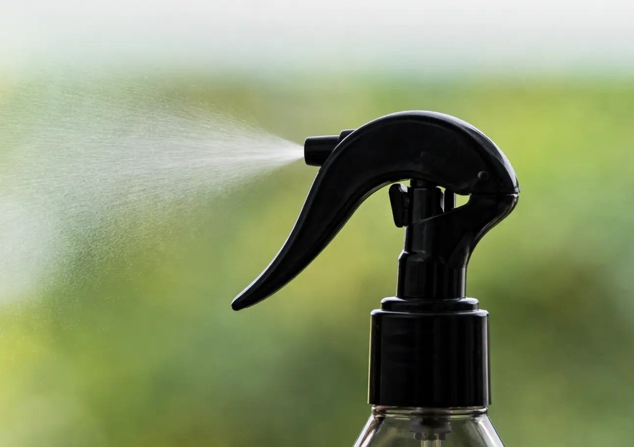 Sort forstøverkapsel på en gennemsigtig flaske, der sprayer en fin tåge mod en sløret grøn baggrund.