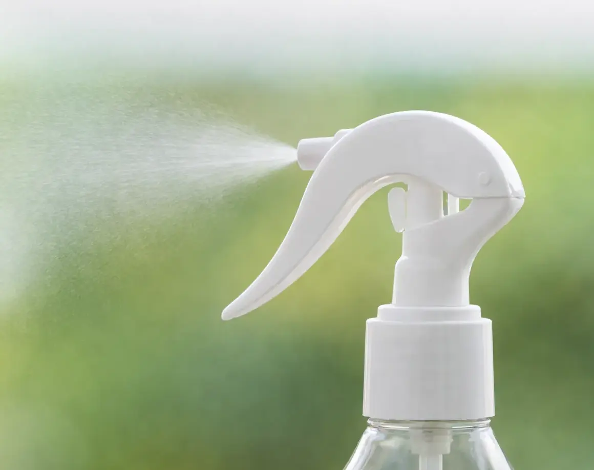 Hvid sprøjtehåndtag på en gennemsigtig flaske, der spreder en fin mist med en grøn, sløret baggrund.
