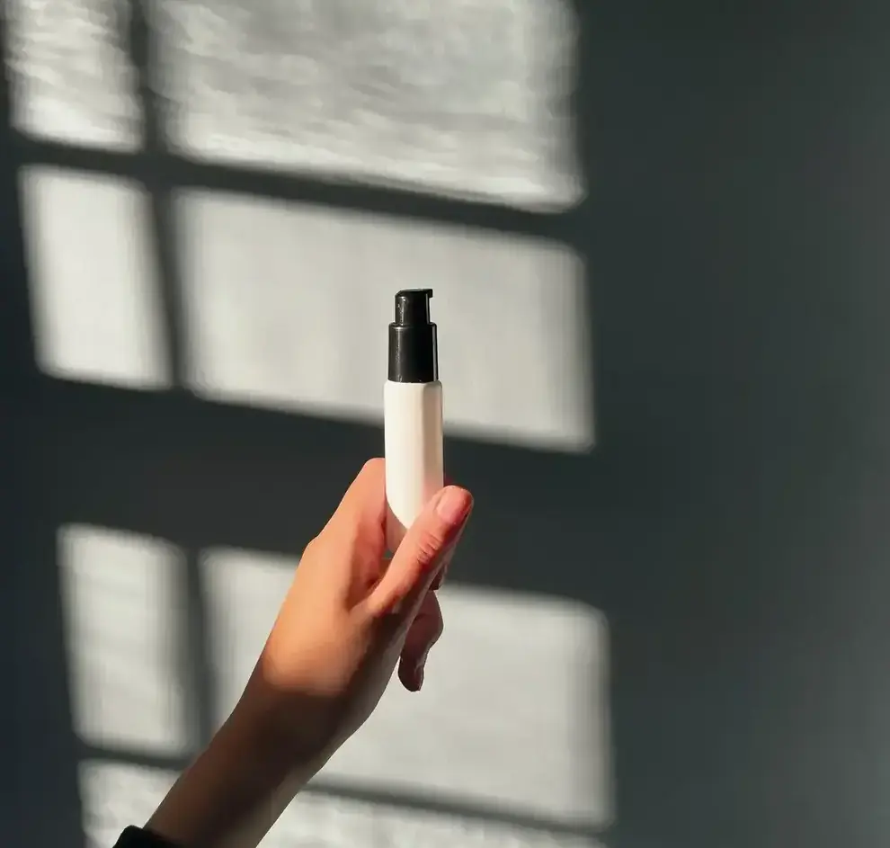 Hånd holder en hvid kosmetisk pumpeflaske med sort låg mod en væg med vinduesskygger.