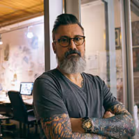 Bearded man with glasses and tattooed arms standing with crossed arms in an office.