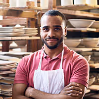 Confident man wearing a white apron and pink polo shirt standing with arms crossed in a pottery workshop with shelves of clay pots behind him.