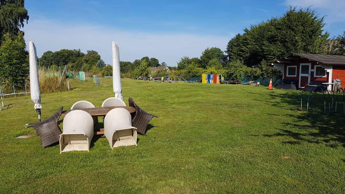Leerer Garten mit umgestürzten Stühlen um Holztisch, Sonnenschirmen und Spielplatz im Hintergrund.