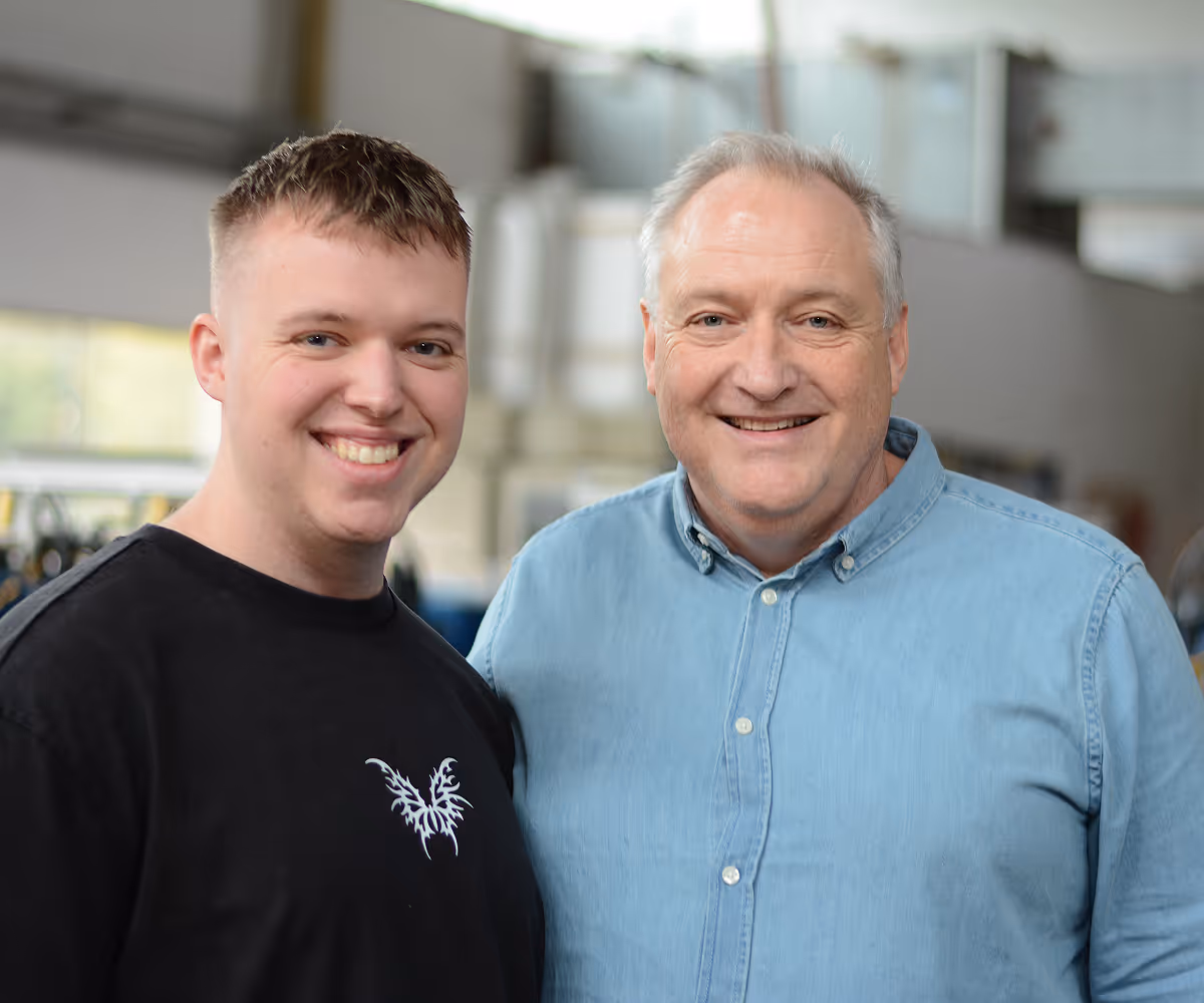 Zwei lächelnde Männer, einer in einem schwarzen T-Shirt und der andere in einem hellblauen Hemd, stehen nebeneinander in einer hellen Innenraumumgebung.