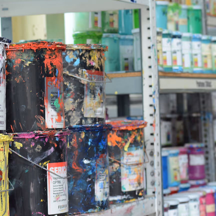 Mehrere Farbdosen mit bunten Farbspritzern stehen gestapelt auf einem Regal in einem Lagerraum.