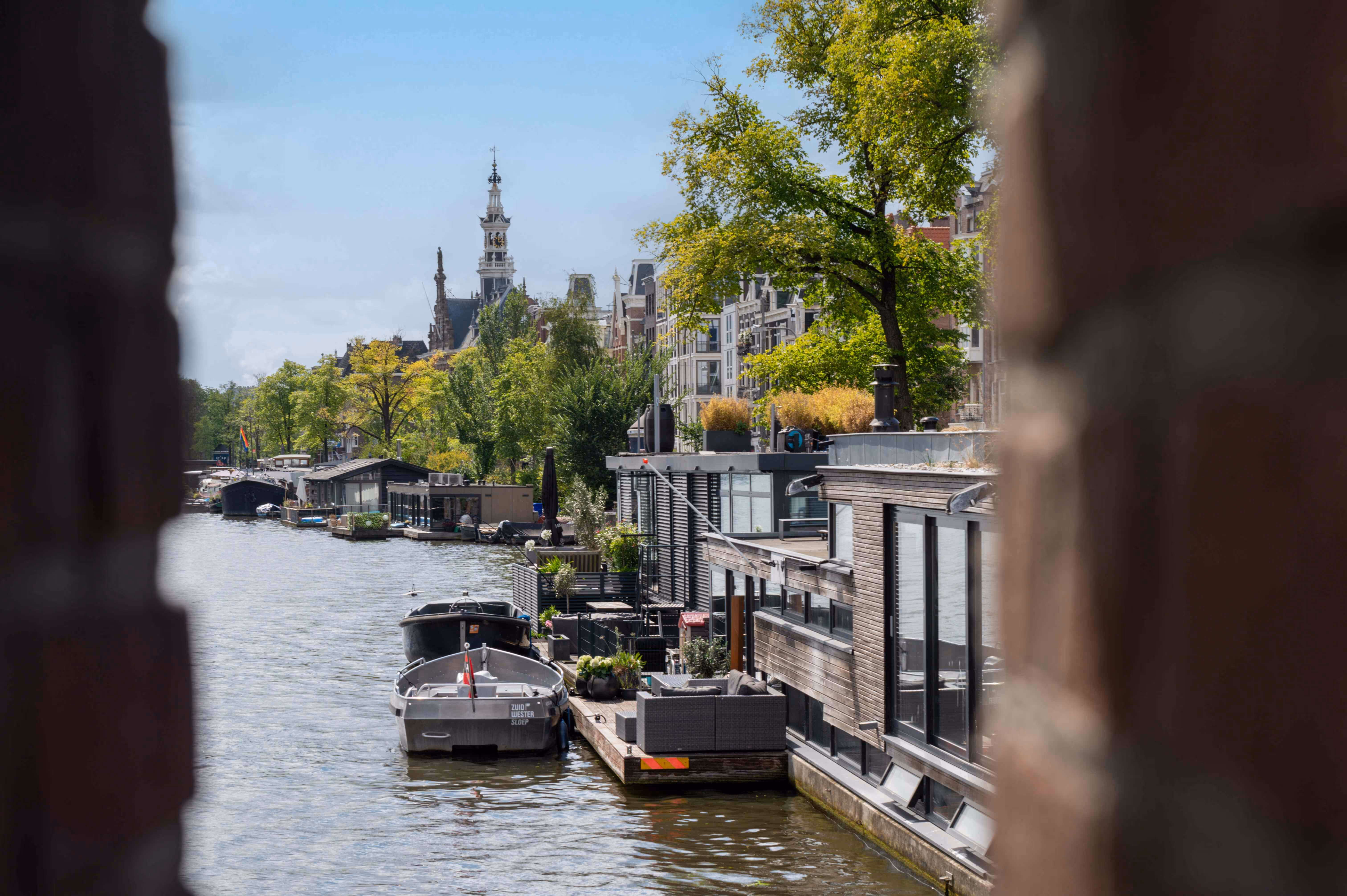 Canal in Amsterdam with houseboats, trees, and historic buildings including a church tower under a clear sky.